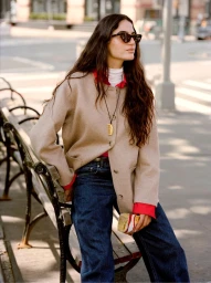 Model wearing a tan jacket, red collar, and blue jeans seated on a park bench outdoors