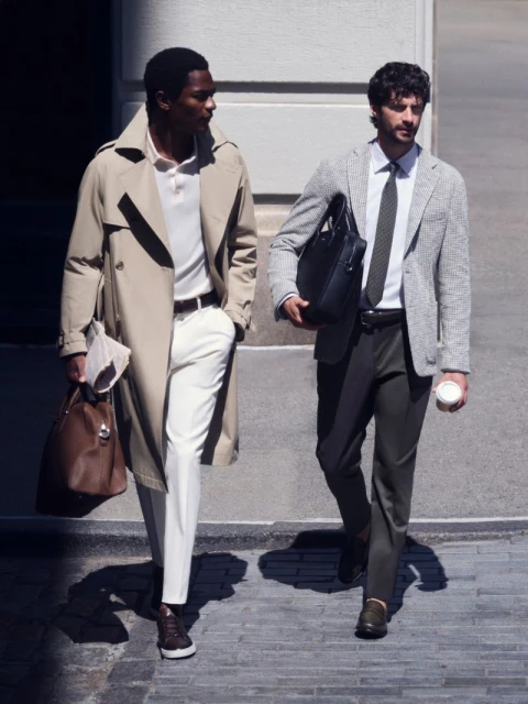 Two men walking outdoors in urban menswear, one in a beige trench coat and white trousers, the other in a grey blazer
