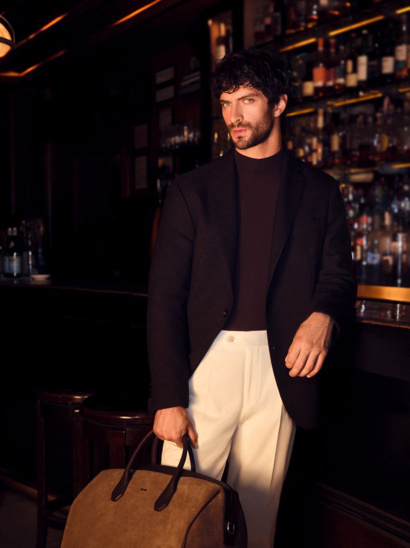 Man wearing dark blazer and white trousers holding a brown leather holdall bag inside a bar
