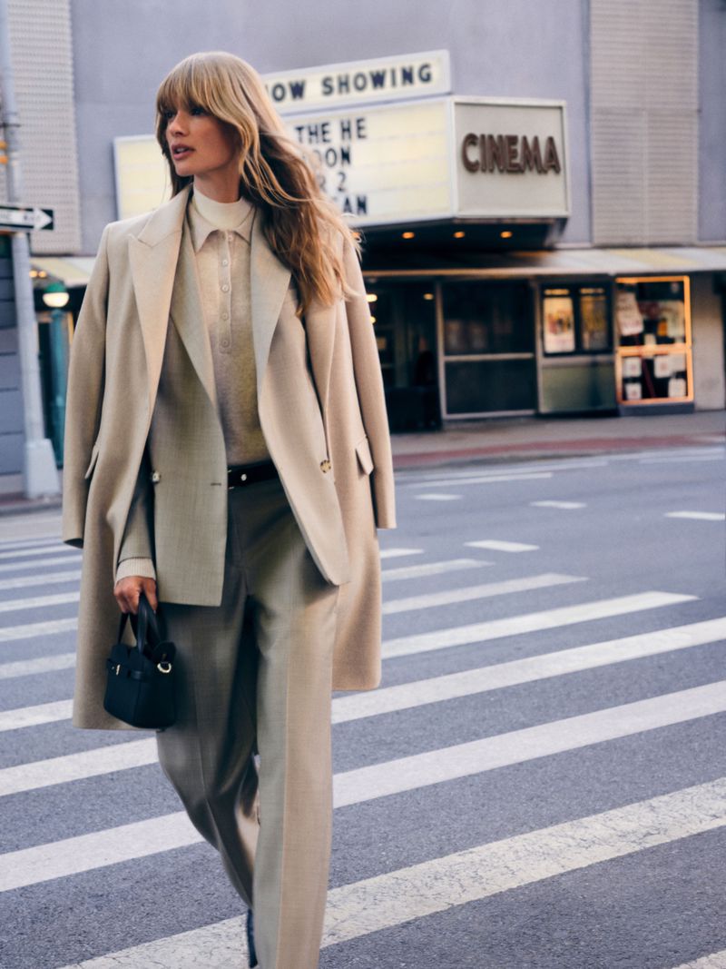 Model wearing a neutral beige suit and matching coat walking across a city street in front of a cinema building.