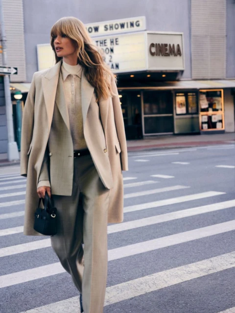 Model wearing a neutral beige suit and matching coat walking across a city street in front of a cinema building.