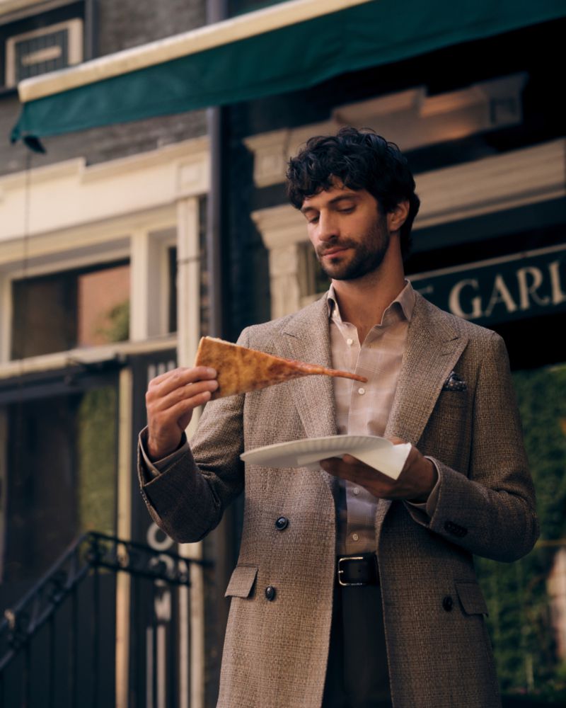 Man in a double-breasted tweed blazer holding a slice of pizza outdoors in front of a building.