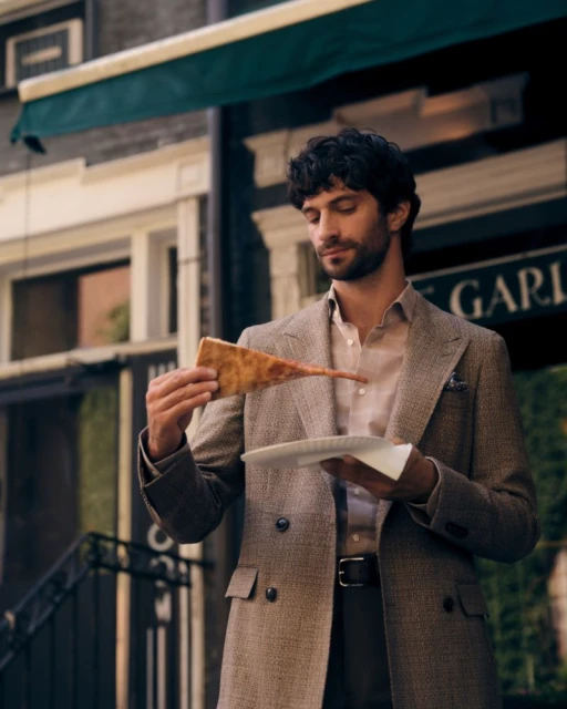 Man in a double-breasted tweed blazer holding a slice of pizza outdoors in front of a building.