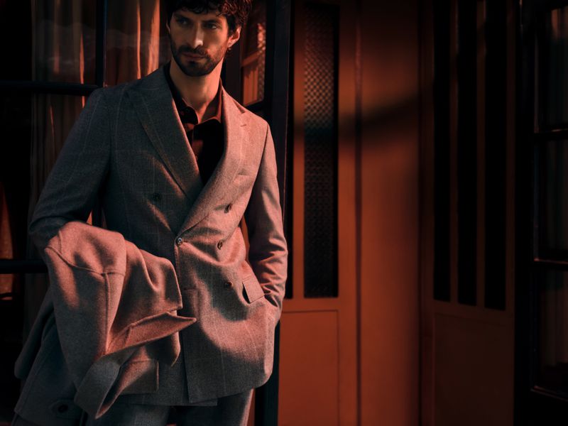 Man wearing a grey double-breasted suit holding a jacket in a dimly lit interior with warm lighting.