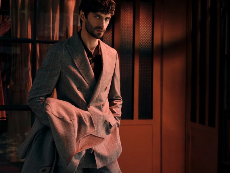 Man wearing a tailored grey double-breasted suit holding a coat in a warmly lit interior doorway.