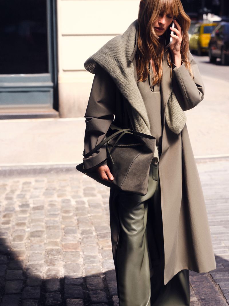 Model in olive green coat and trousers holding a large tote bag while walking on a city street