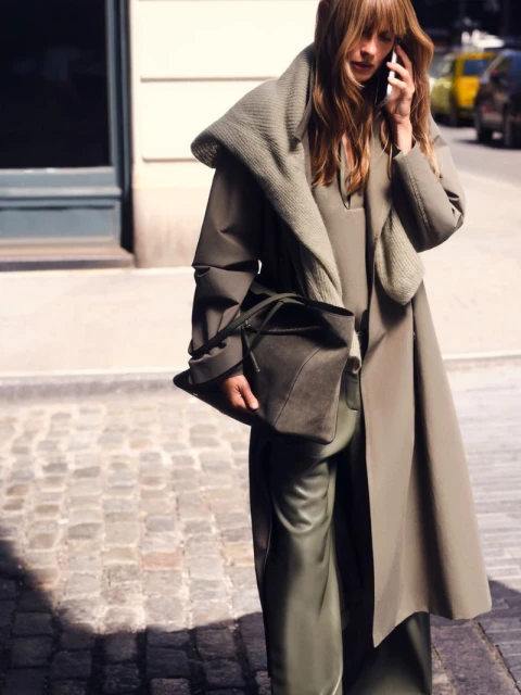 Model in olive green coat and trousers holding a large tote bag while walking on a city street