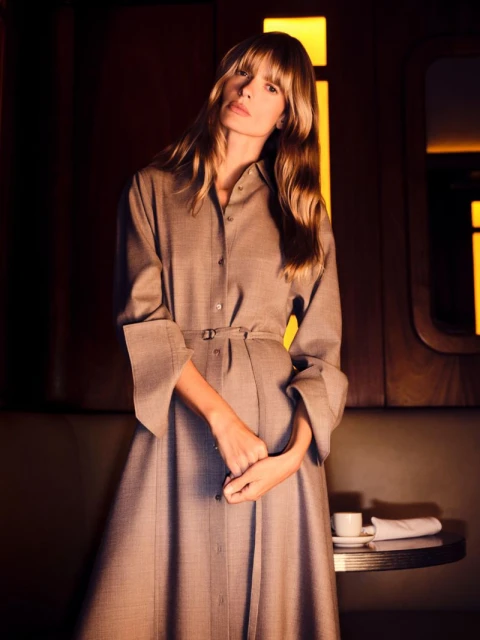 Model in a brown belted shirt dress standing inside a dimly lit interior setting with warm lighting.