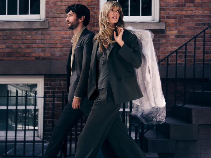 Two models walking outside a brick building, wearing a tailored suit and an olive green blazer with trousers carrying a garment bag.