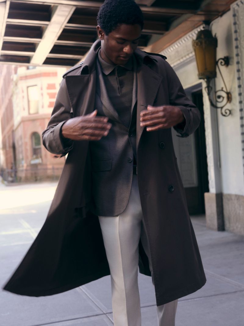 Black man in a brown trench coat and light trousers walking down an urban street