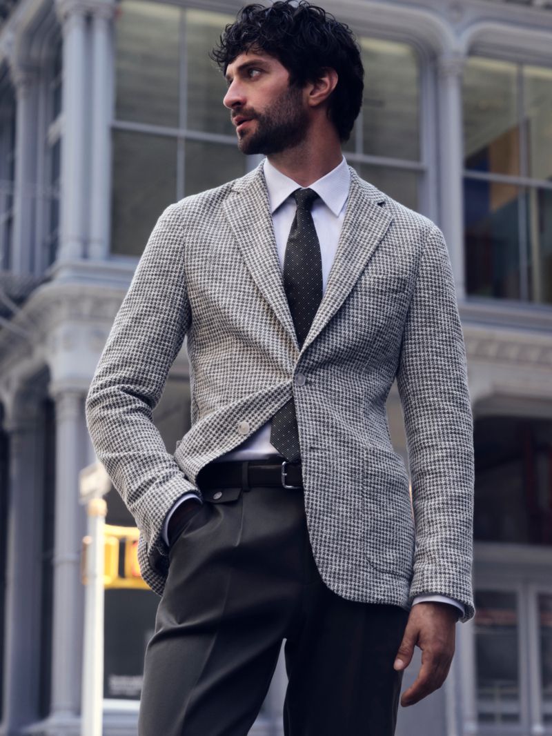 Man wearing a grey tweed blazer and dark tie standing in front of classical stone architecture.