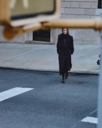Model in a long dark coat walking across a city street viewed past a blurred street sign arm