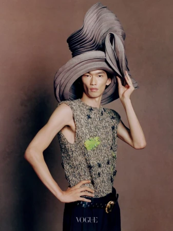 Model wearing a large sculptural grey hat and textured sleeveless top against a warm background for Vogue China Men