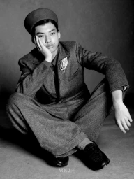 Model seated on the floor wearing a tailored tweed suit and cap with hand supporting chin, photographed in monochrome