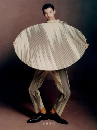 Model holding a large circular pleated garment wearing matching trousers and black shoes against a dark studio backdrop