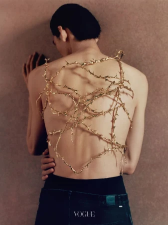 Shirtless model viewed from behind with a gold thorny vine sculpture draped across their back against a textured wall.