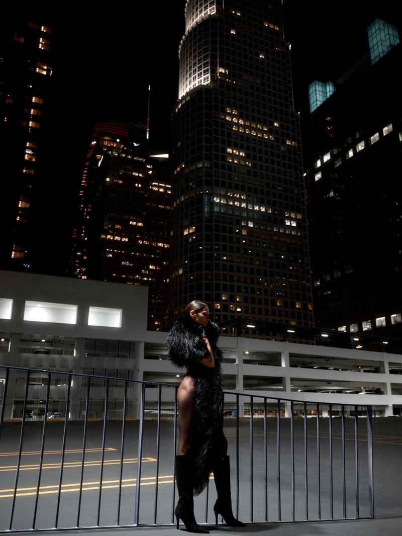 Model standing on a rooftop at night wearing a dark feathered coat and tall black boots against illuminated skyscrapers