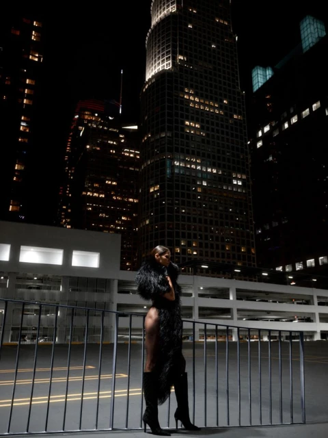 Model standing on a rooftop at night wearing a dark feathered coat and tall black boots against illuminated skyscrapers