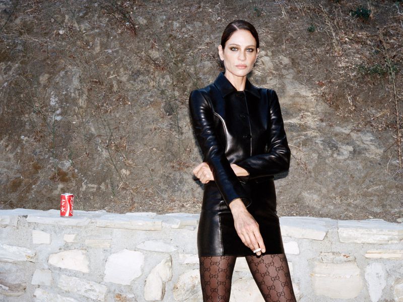 Model Amber Valletta wearing a black leather jacket and matching skirt standing in front of a stone wall