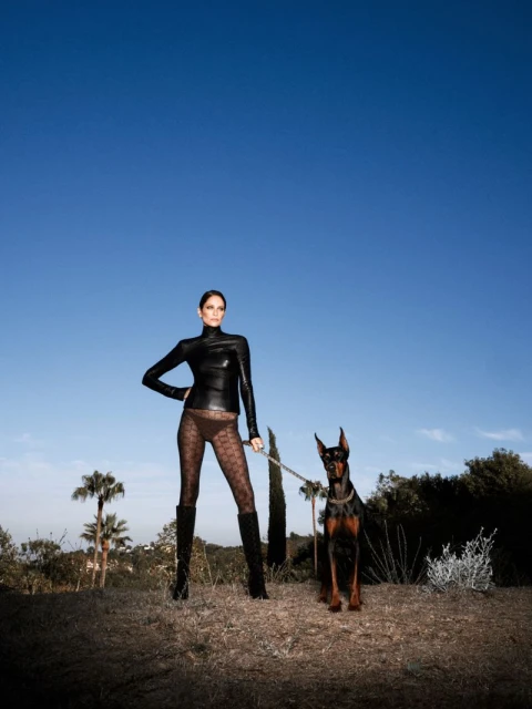 Model Amber Valletta wearing black turtleneck and patterned tights holding Doberman leash under clear blue sky