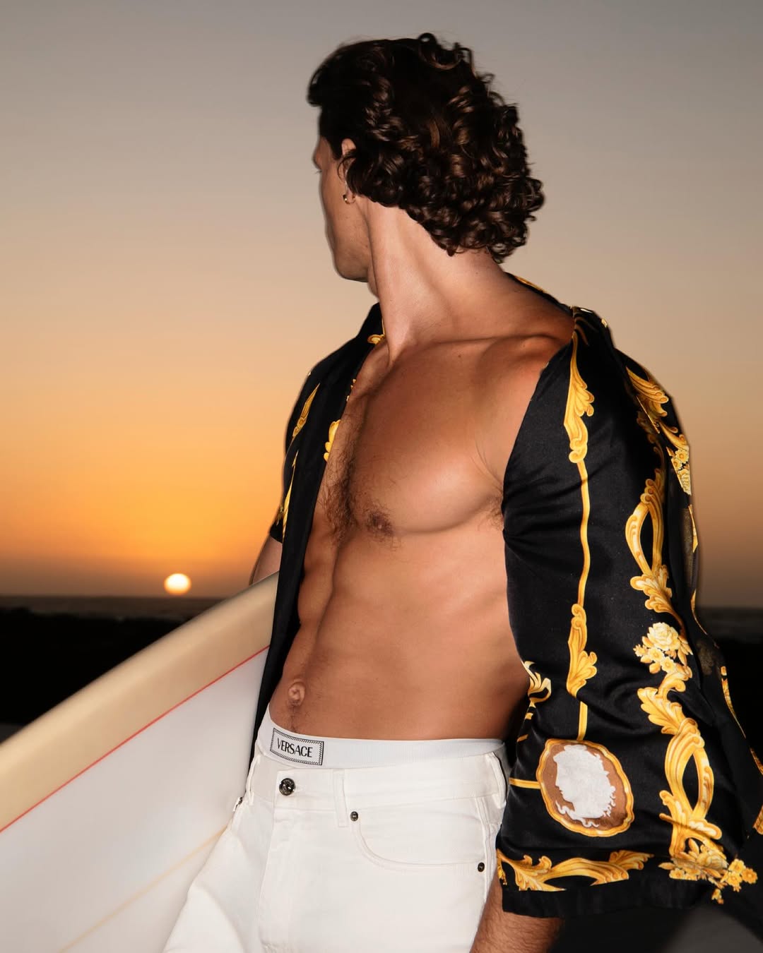 Shirtless man with a surfboard stands at the beach at sunset. He wears white jeans and an unbuttoned Versace shirt.