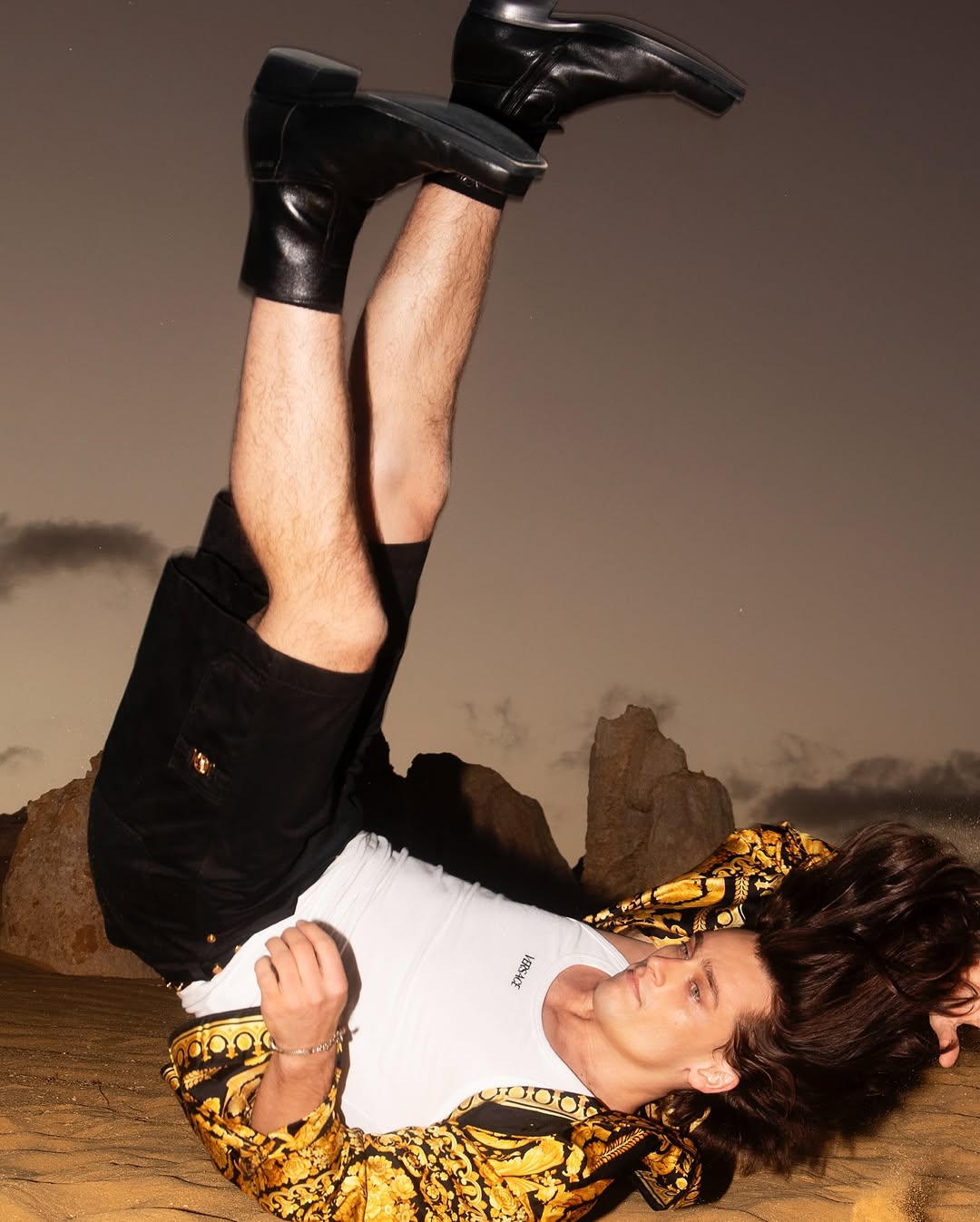 Man lying upside down on sand wearing a black patterned shirt, white tank top, and black leather boots