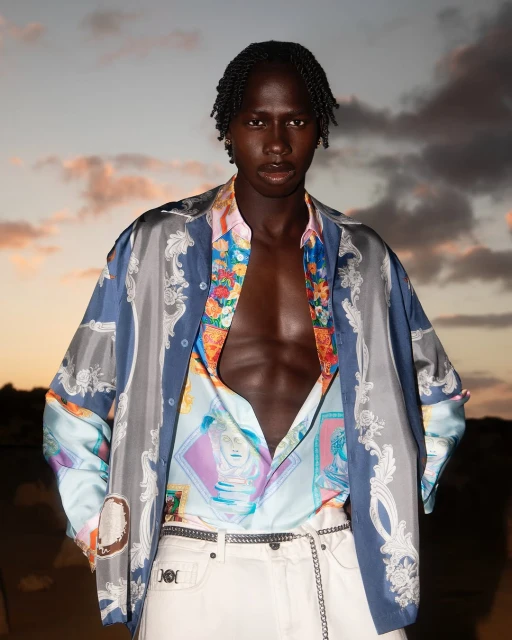 Portrait of a black male fashion model wearing an open patterned shirt and white pants, posing against a sunset sky background.