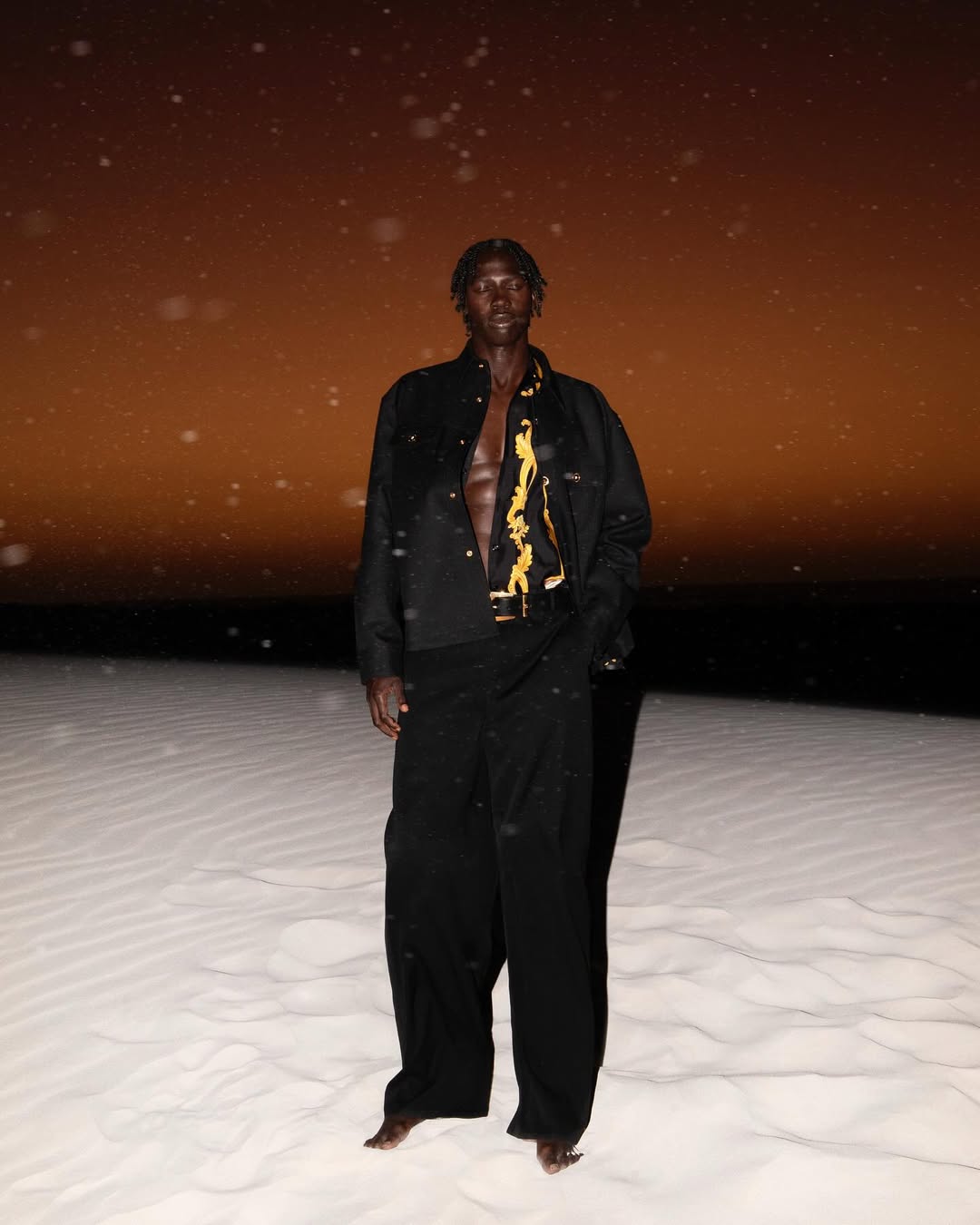 Dark-skinned male model in an open black shirt and loose pants poses on a white sand dune at night with a golden sky.