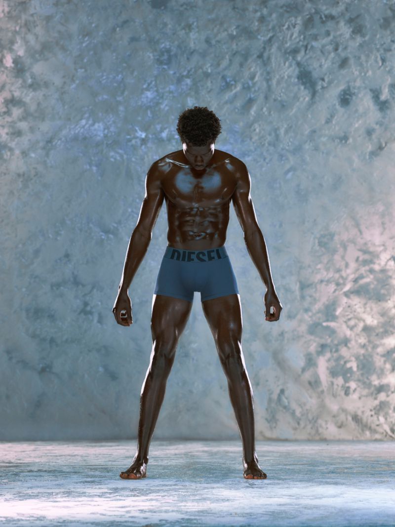Athletic, muscular Black male stands with open posture against a textured gray wall wearing blue Diesel underwear in studio lighting.