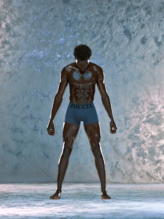Athletic, muscular Black male stands with open posture against a textured gray wall wearing blue Diesel underwear in studio lighting.