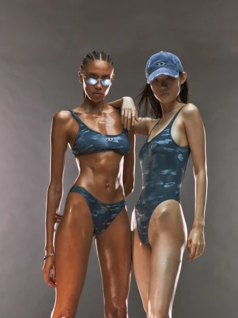 Two female models posing in matching blue printed swimwear, sunglasses, and a cap against a neutral studio backdrop.