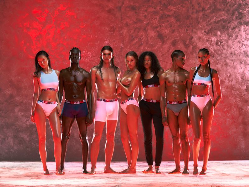 Seven people pose in a studio wearing Diesel branded underwear against a red backdrop with soft lighting for a fashion advertisement.