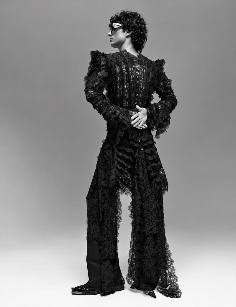 Model wearing a black lace patterned suit and sunglasses standing against a seamless grey background