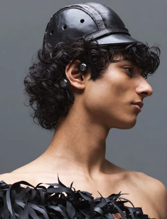 Close-up profile of a male model wearing a dark leather cap and textured black garment against a grey background.