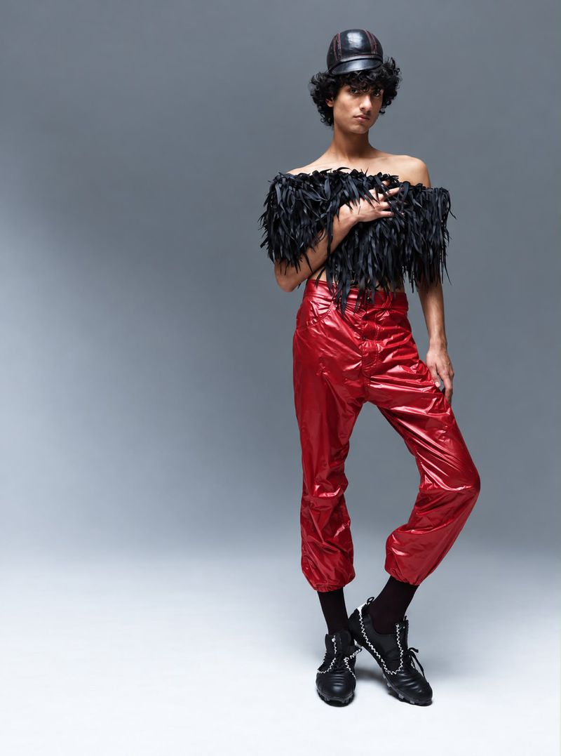 Model Max Everist wearing a black feather top and red trousers against a grey studio backdrop for The Hunger Magazine