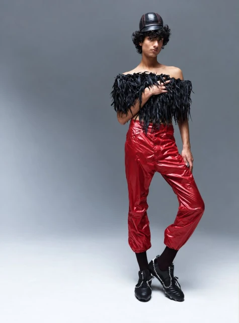 Model Max Everist wearing a black feather top and red trousers against a grey studio backdrop for The Hunger Magazine