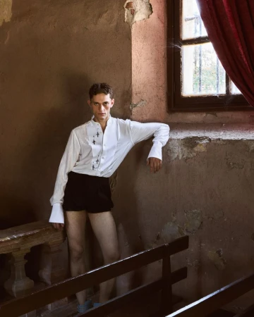 Model leaning against a weathered stone wall in a white shirt and black shorts with a red curtain by a window