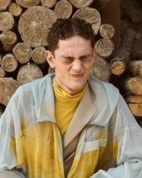 Close-up portrait of a male model with eyes closed tightly, wearing a translucent white jacket over a yellow turtleneck sweater.