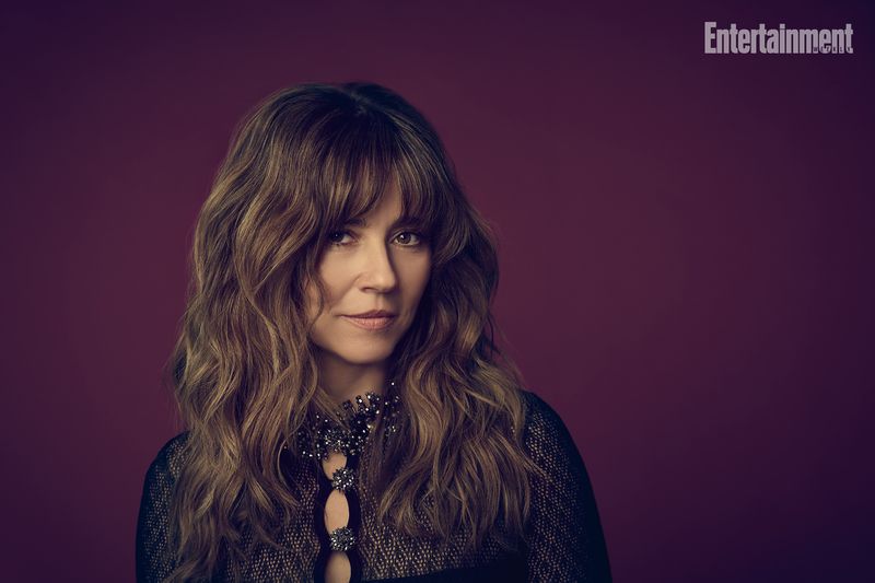 Portrait of Linda Cardellini with wavy brown hair wearing a dark textured top against a solid dark plum background