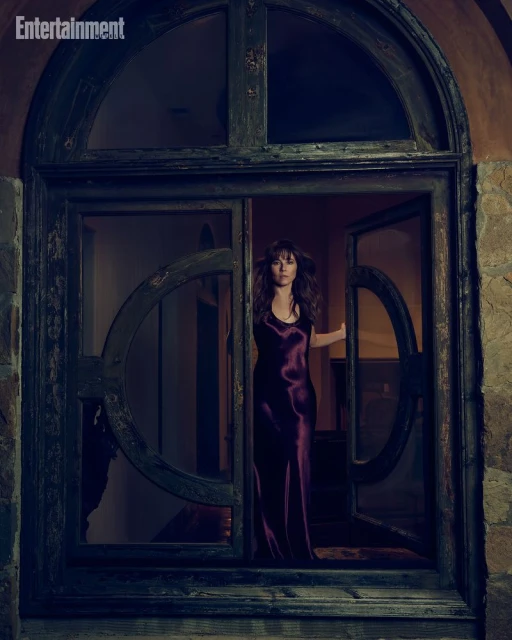 Linda Cardellini wearing a dark purple satin slip dress standing in an ornate arched wooden doorway.