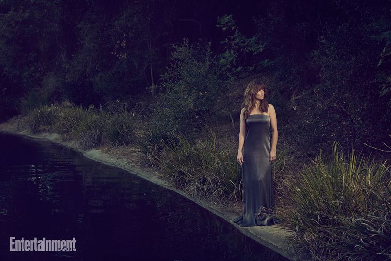 Linda Cardellini standing by a dark body of water in a long grey gown near dense trees and foliage