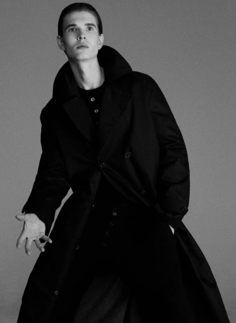 Model in a black trench coat posing against a grey seamless background for a Wallpaper Magazine editorial shoot.