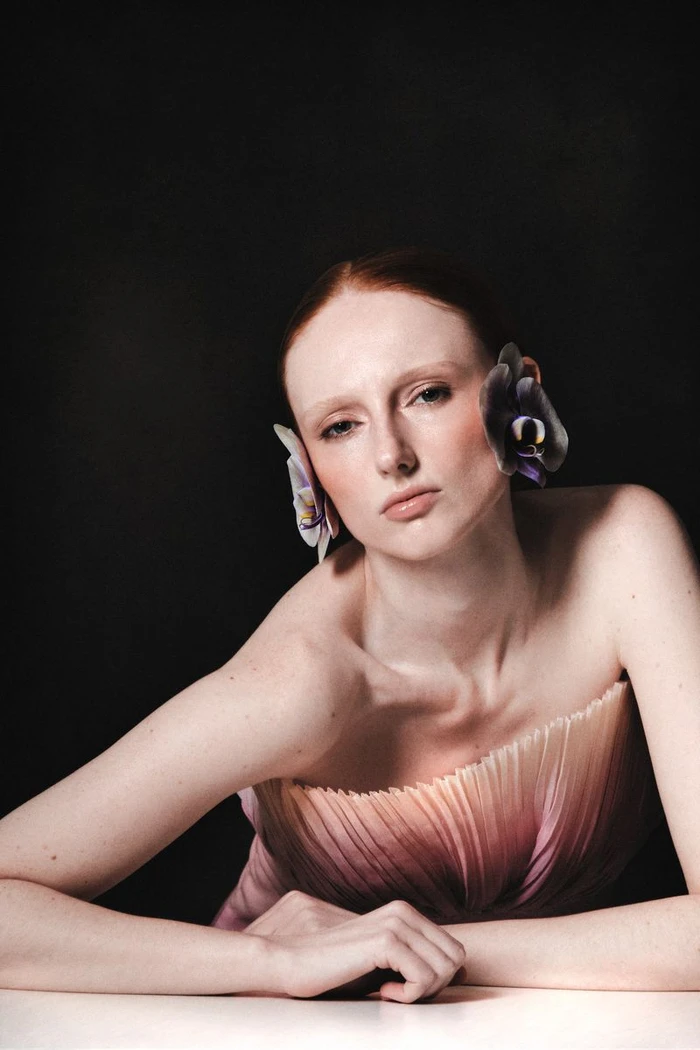 Model with red hair and orchids on ears wearing a gradient pink and white strapless dress against a black background.