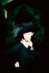Woman in black feathered hat and black long-sleeve top posing outdoors at night in front of a dark stone archway