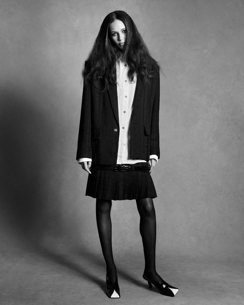 Model Binx Walton wearing a black blazer, white shirt, and pleated skirt against a plain studio background.