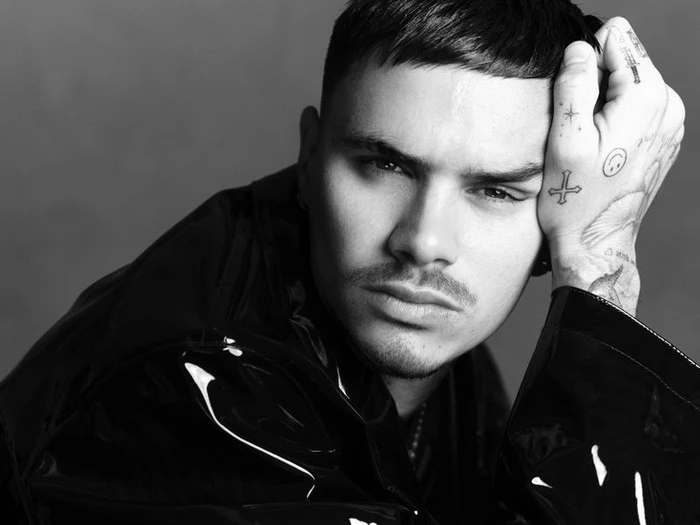 Close-up monochrome portrait of Romeo Beckham with hand to head showing tattoos, wearing shiny black outerwear for Double Vision.