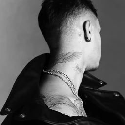 Close-up monochrome portrait of Romeo Beckham showing neck and shoulder tattoos, silver chains, and a black leather jacket.