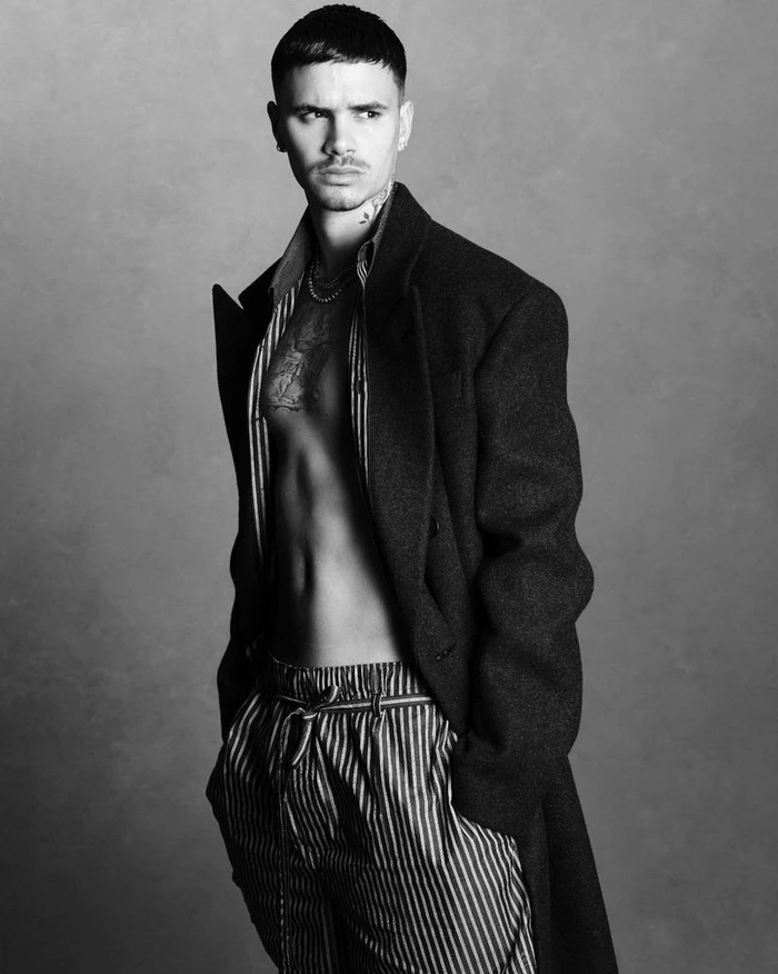Romeo Beckham wearing an open dark coat and striped trousers against a grey backdrop