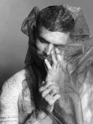 Monochrome portrait of Romeo Beckham draped in sheer dark fabric with visible tattoos on arm and hand.