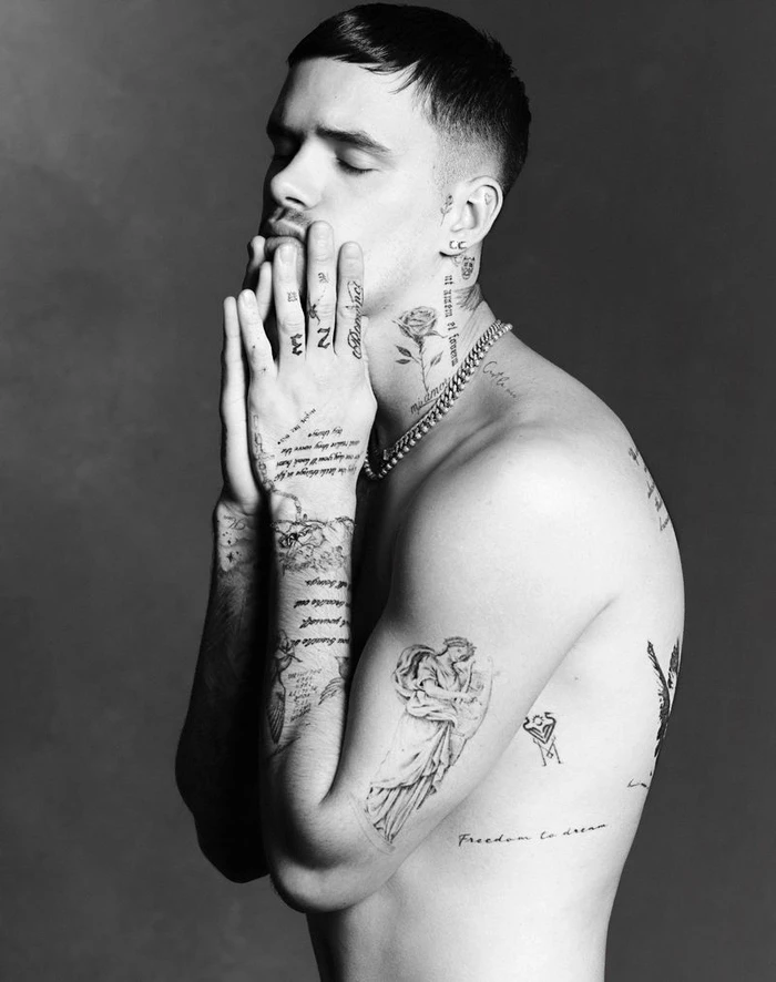 Portrait of Romeo Beckham in profile with hands to face featuring tattoos and silver chain necklace on grey seamless background